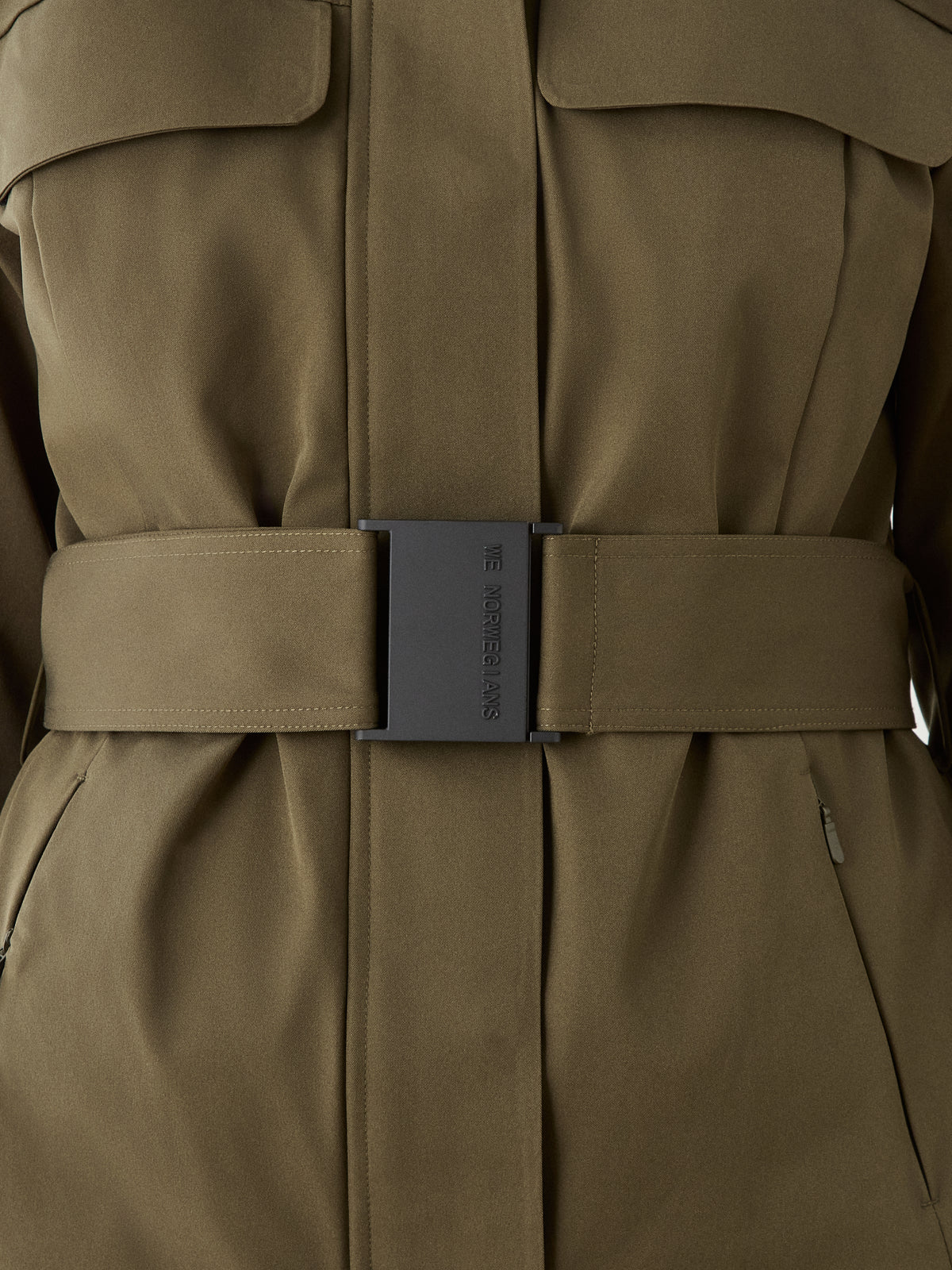 Close-up of the olive green Finse Ski Jacket Women by We Norwegians, featuring a wide belt with a large rectangular “We Are Versus” buckle and sharp, modern ski jacket details.