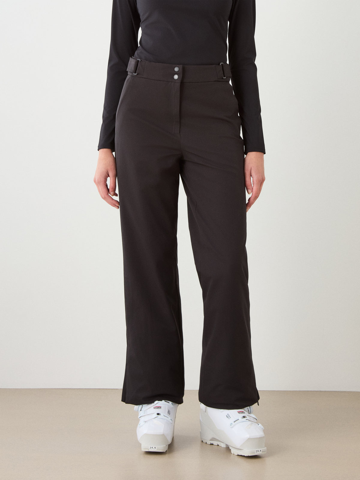 A person in black We Norwegians Finse Ski Pant Women, a black long-sleeve top, and white ski boots stands against a plain background, shown from neck to feet.