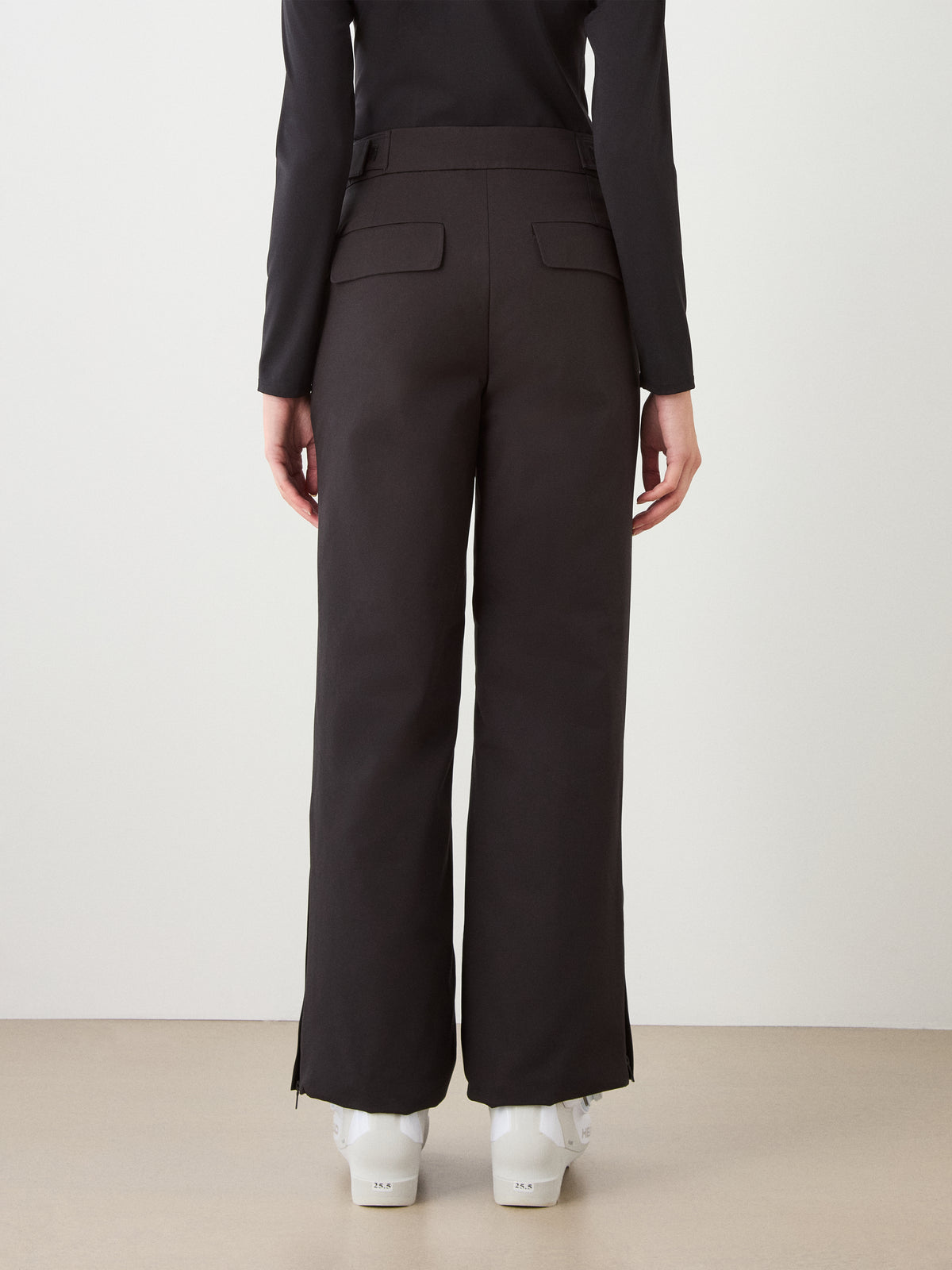 A person faces away in We Norwegians Finse Ski Pant Women—black wide-leg insulated ski pants with back flap pockets—paired with a black top and white shoes, striking a perfect alpine look against a light background.