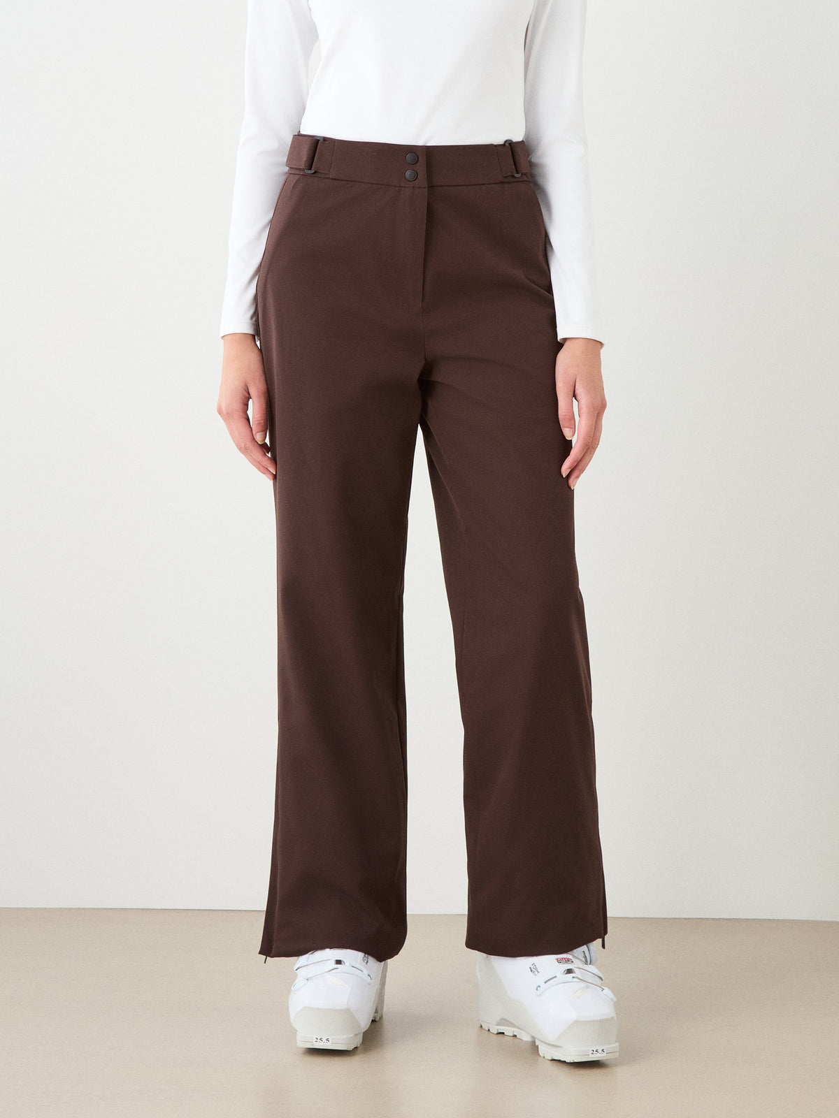Wearing a white long-sleeve top, We Norwegians Finse Ski Pant Women in brown straight-leg style, and white boots, a person stands against a plain background—only visible from the shoulders down.