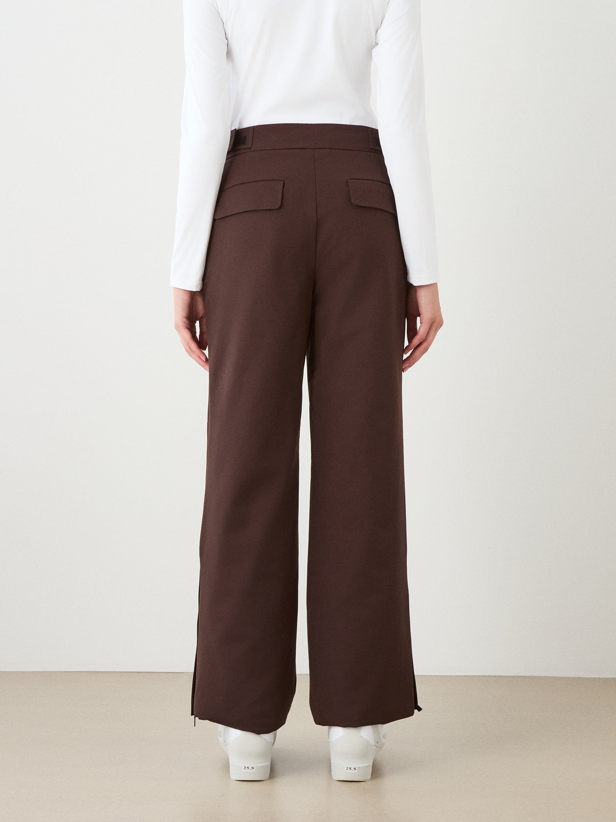 A person stands with their back to the camera, wearing We Norwegians Finse Ski Pant Women in brown, a long-sleeve white top, and white shoes against a plain background.