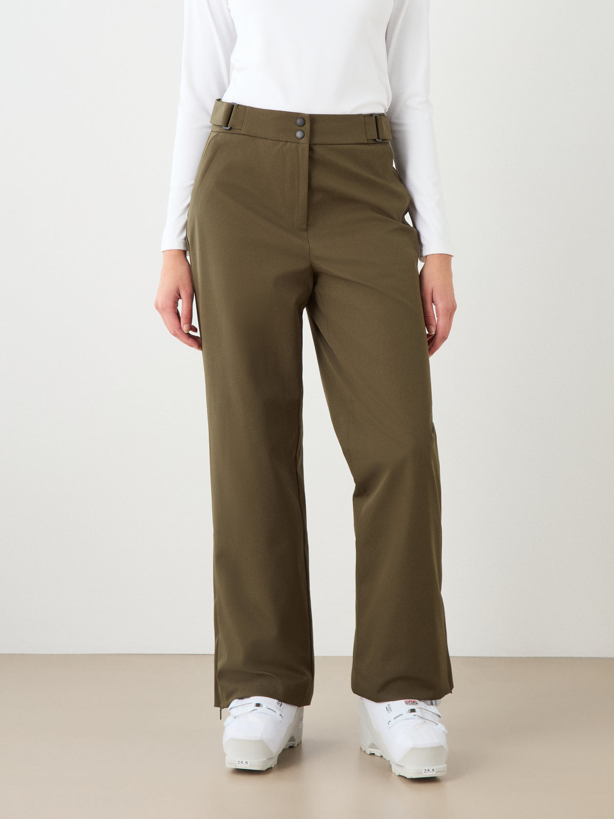 Person wearing a white long-sleeve top, We Norwegians Finse Ski Pant Women in olive green, and white sneakers, standing against a plain light background with hands relaxed by their sides.