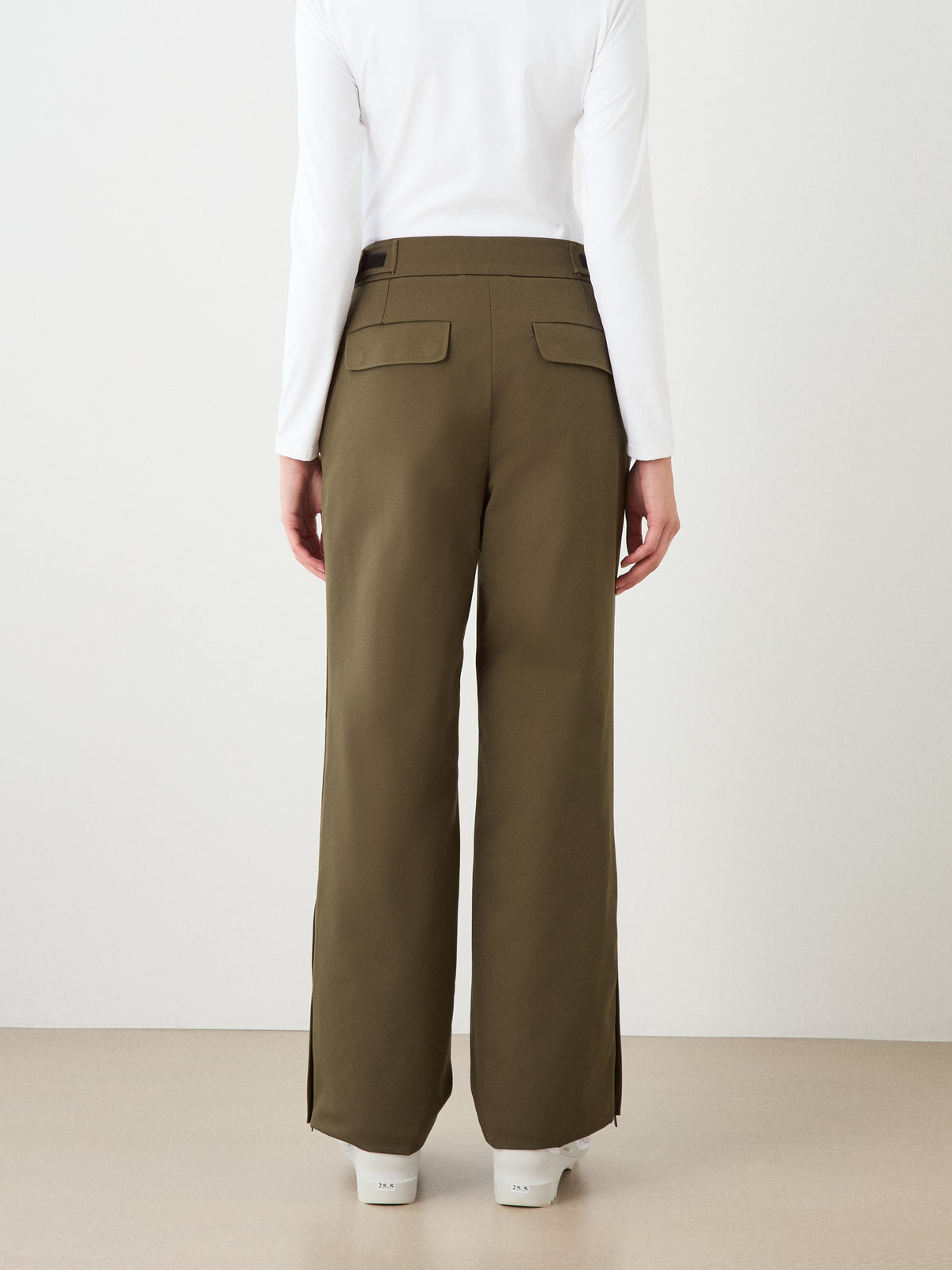 Person stands with their back to the camera, wearing We Norwegians Finse Ski Pant Women in olive green, a white long-sleeve top, and white shoes against a plain light background.