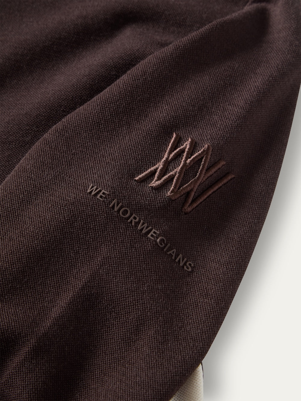 Close-up of a dark brown We Norwegians Voss ZipUp Women sleeve featuring an embroidered logo above embossed text.