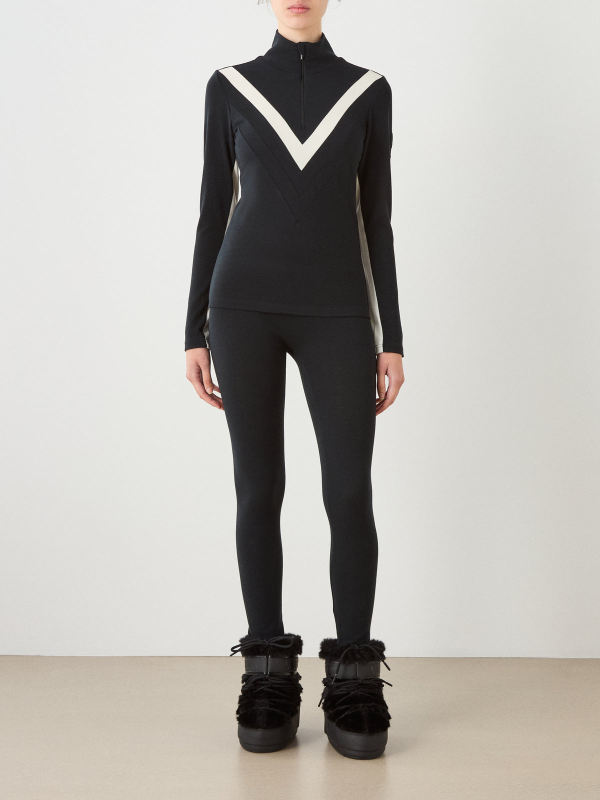 A person wearing a black We Norwegians Voss Chevron ZipUp women’s top with a high collar, paired with matching black leggings and black faux-fur lined boots for an après-ski look.