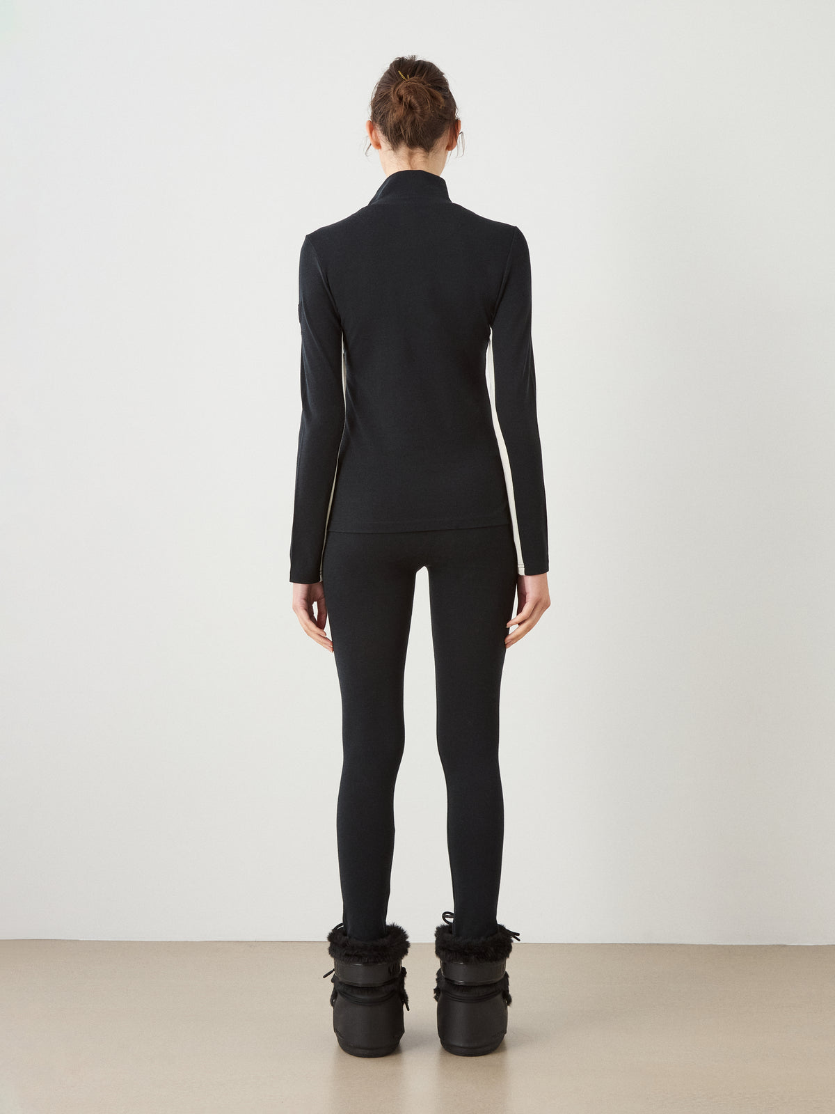 Back view of a person wearing a black We Norwegians Voss Chevron ZipUp Women, matching black leggings, and black boots with faux-fur cuffs.