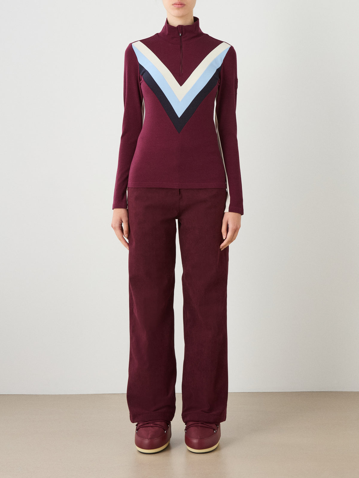 A person wearing a burgundy We Norwegians Voss Chevron ZipUp Women with a light blue, white, and navy pattern across the chest, paired with matching trousers and slip-on shoes.
