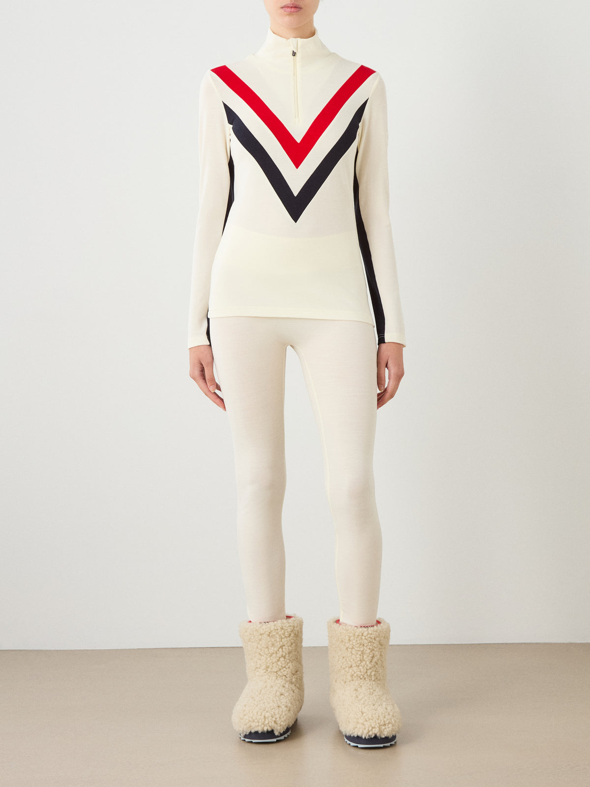 A woman wears a cream-colored We Norwegians Voss Chevron ZipUp Women sweater with a red and black chest design, matching cream leggings, and cream-colored fleece-lined boots for an après-ski look.