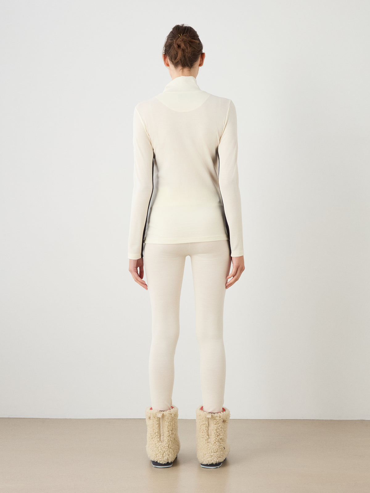 A person seen from the back wearing a cream-colored We Norwegians Voss Chevron ZipUp Women with dark side panels and matching leggings, paired with tan fleece-lined boots for an après-ski look.