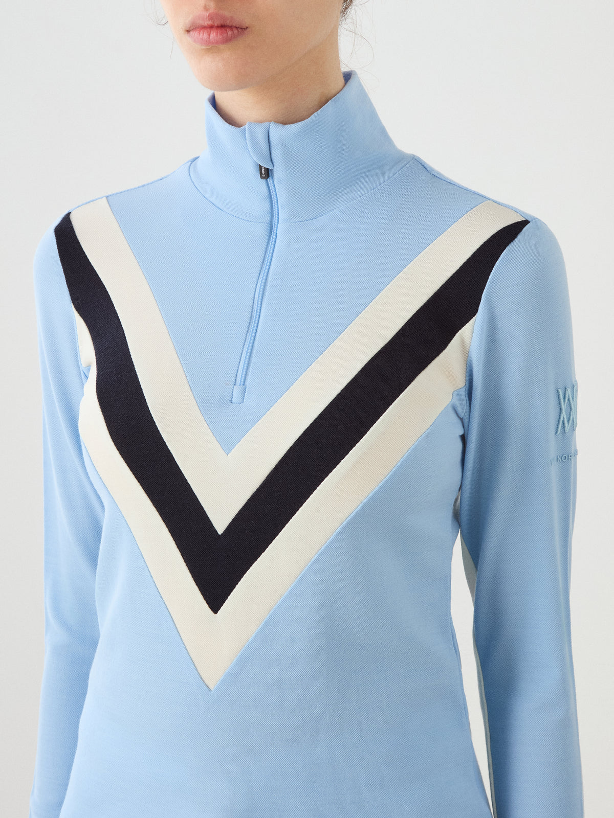 Light blue We Norwegians Voss Chevron ZipUp Women merino wool zip-up mock neck sweater with a white and black chevron stripe design on the chest in a classic après-ski style.