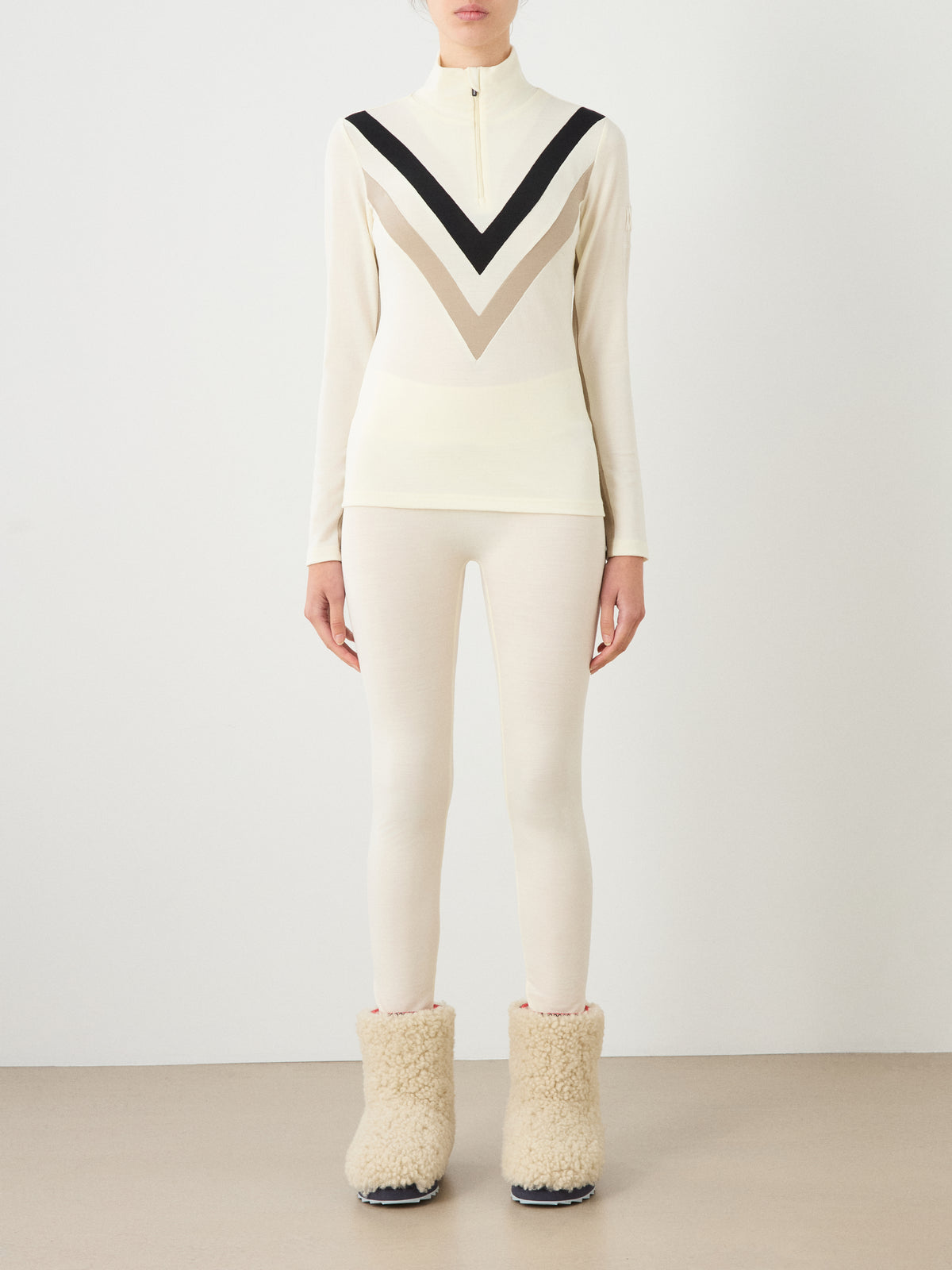 A cream-colored We Norwegians Voss Chevron ZipUp Women merino wool shirt with a black and tan pattern on the chest, paired with matching cream leggings and fuzzy shearling-style boots.