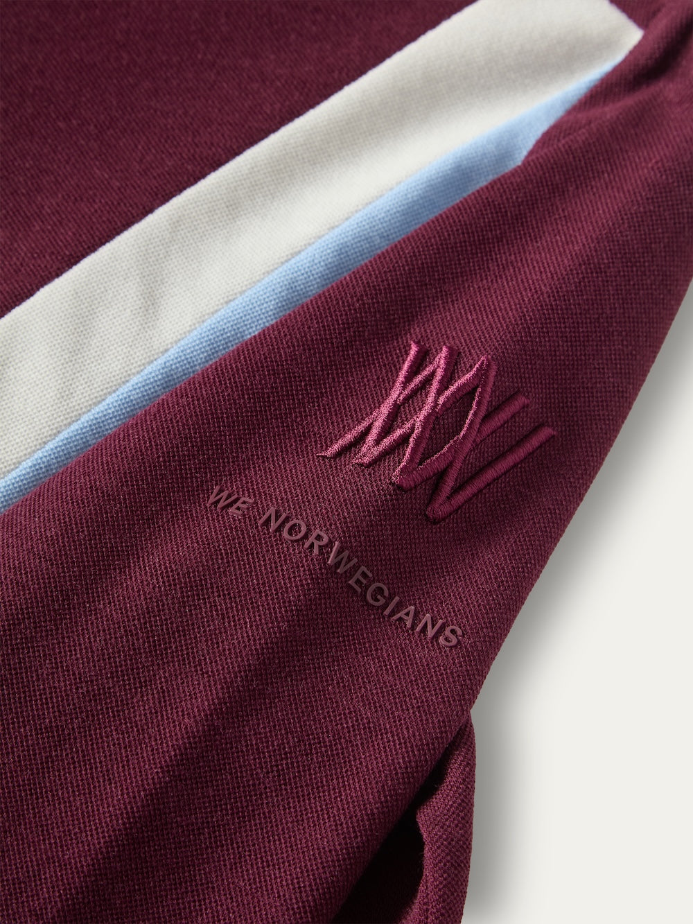 Close-up of a maroon We Norwegians Voss Chevron ZipUp Women long-sleeved top with a white and light blue stripe on the sleeve, featuring an embroidered logo and brand name in matching maroon thread.