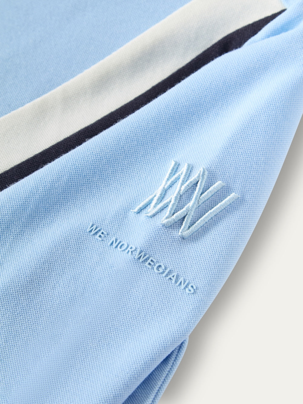 Close-up of a light blue We Norwegians Voss Chevron ZipUp Women sleeve featuring an embroidered logo and brand name, with white and dark blue stripes running along the shoulder.