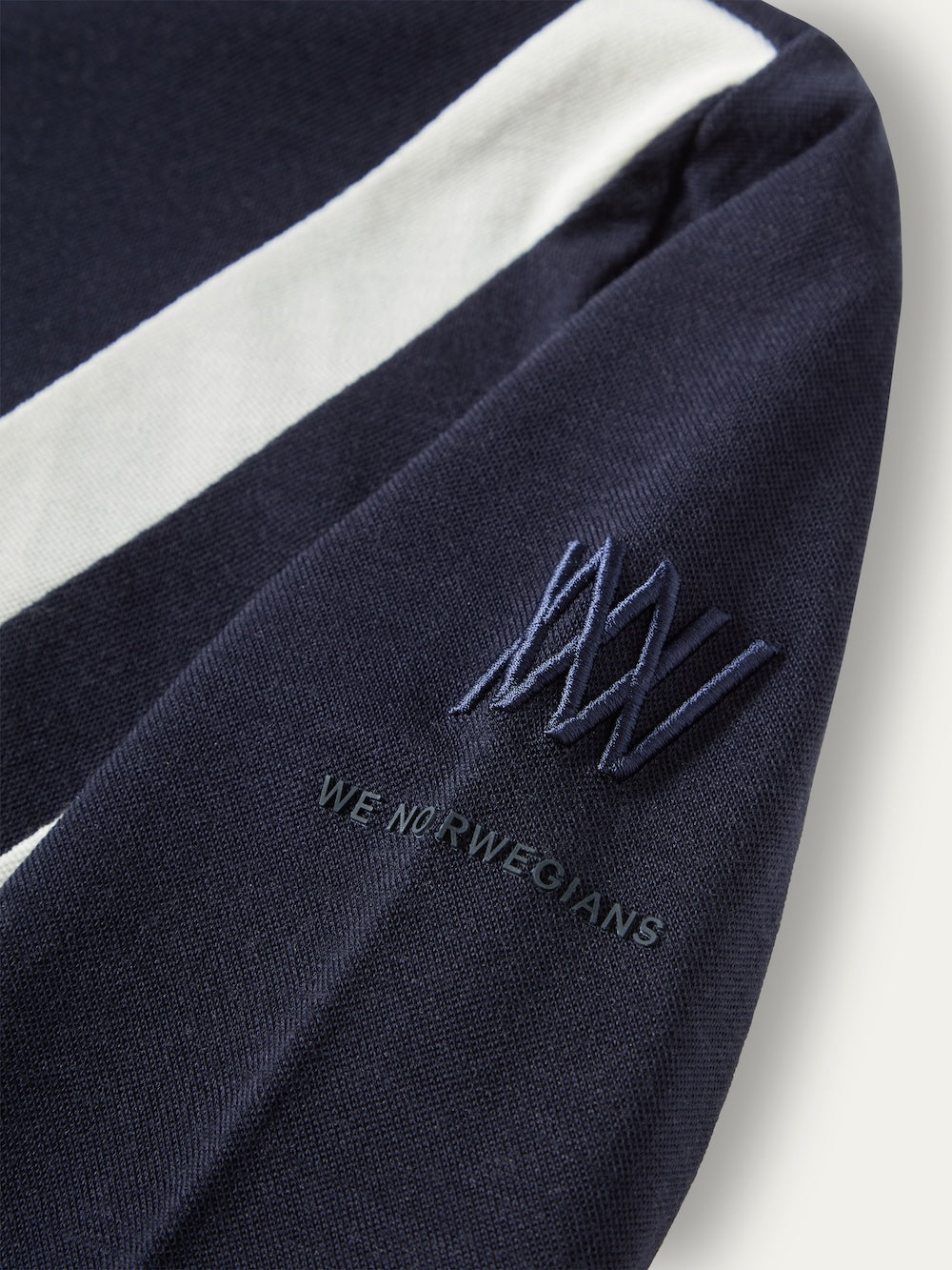 Close-up of a navy blue We Norwegians Voss Chevron ZipUp Women featuring a white stripe down the arm, an embroidered logo, and text on the sleeve.