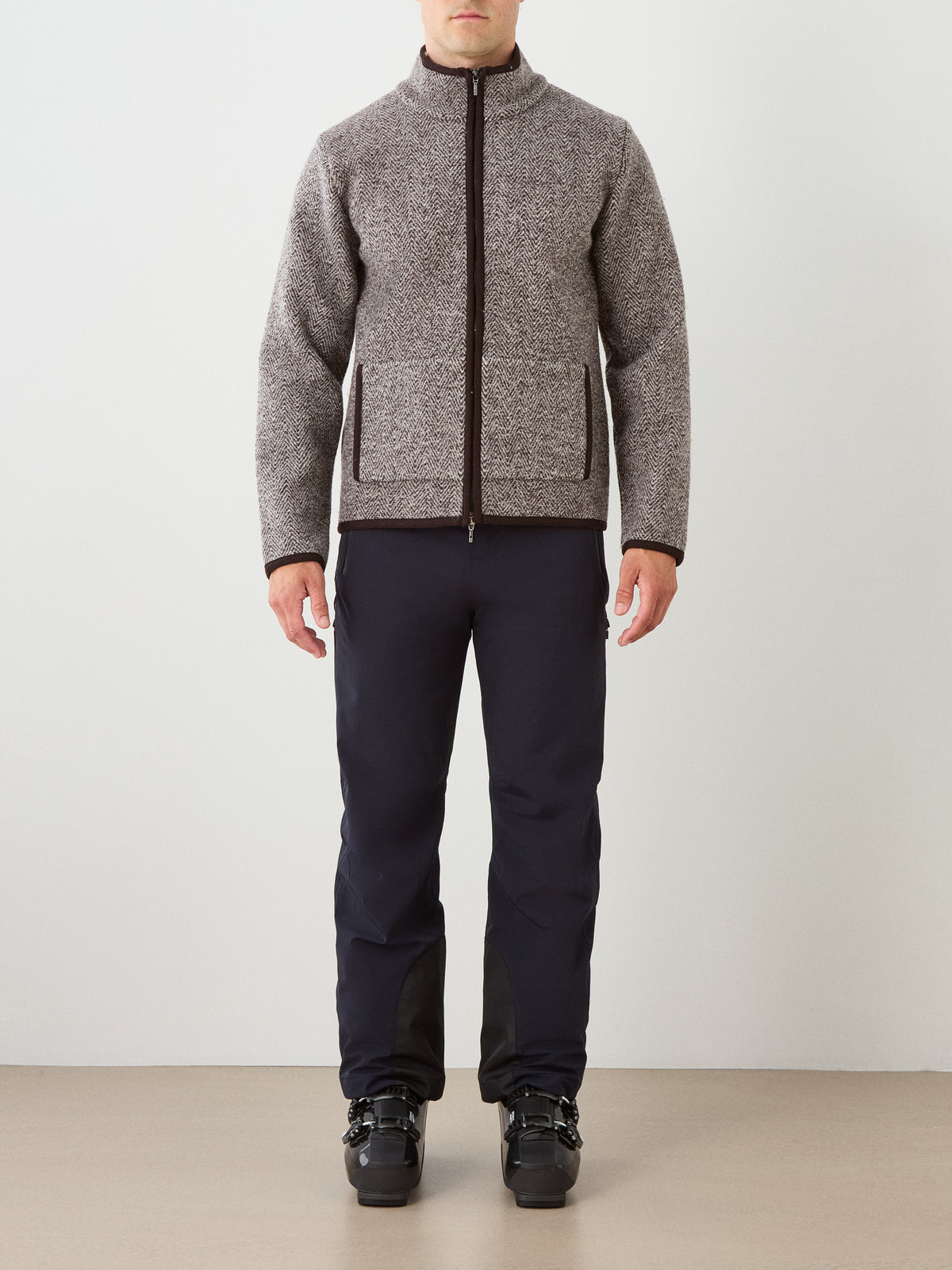 A man stands facing forward against a plain background, wearing the We Norwegians Laerdal Boiled Wool Jacket Men—highlighting Scandinavian craftsmanship—paired with dark pants and black ski boots.