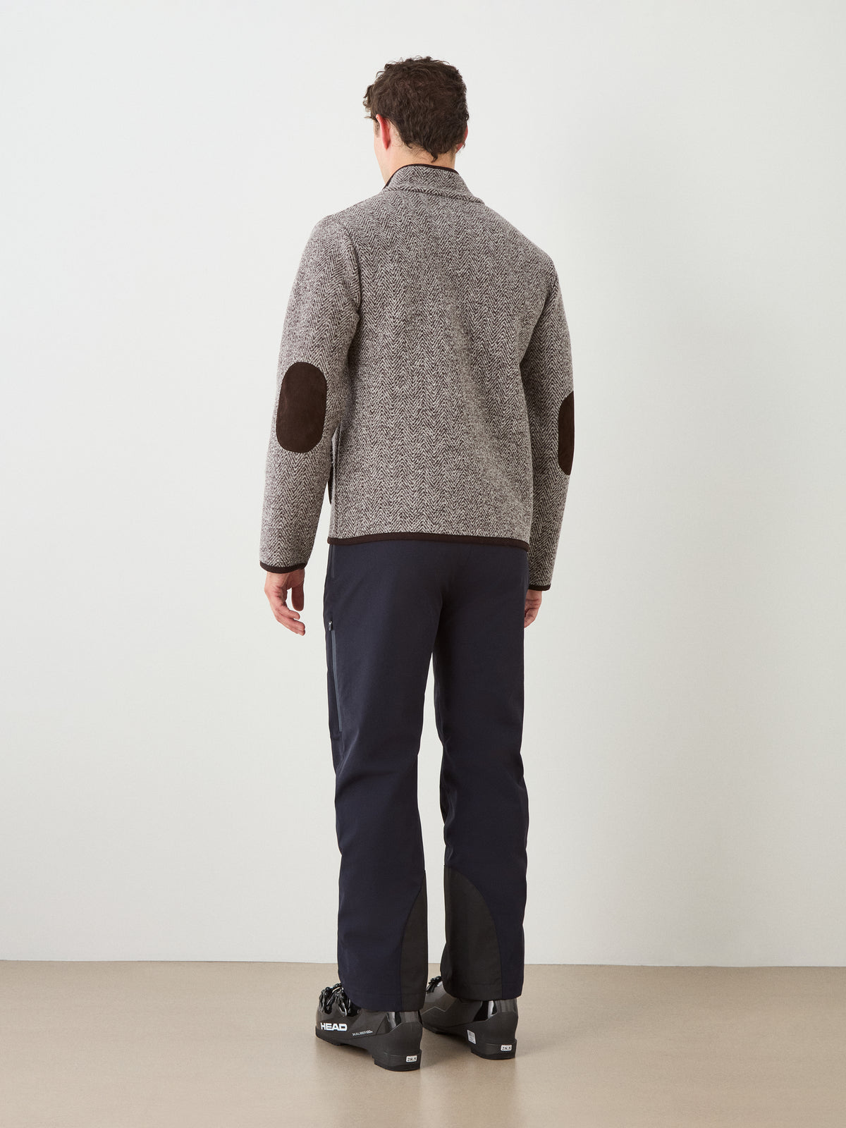 A man stands facing away, wearing the We Norwegians Laerdal Boiled Wool Jacket with brown elbow patches—an embodiment of Scandinavian craftsmanship—paired with navy pants and black shoes, set against a plain light-colored wall and tan floor.