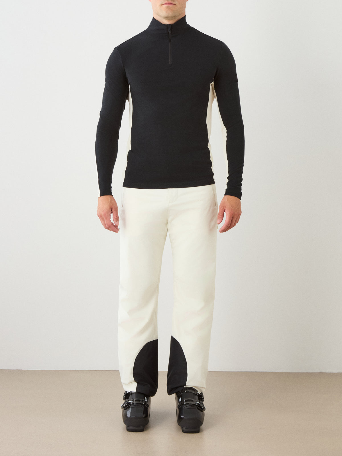 A person stands against a plain background wearing the We Norwegians Voss ZipUp Men, black in color, paired with white pants featuring black ankle patches and black ski boots.
