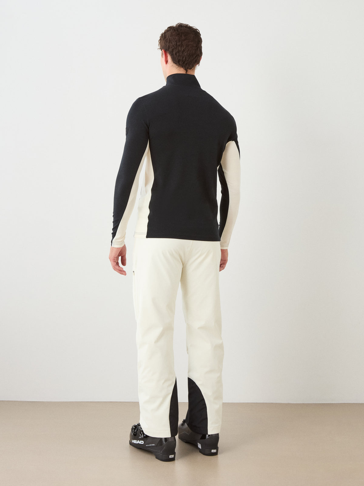 A person stands facing away, wearing the We Norwegians Voss ZipUp Men, a black and white color block activewear long-sleeve top, paired with white pants featuring black patches on the inner calves and black shoes, on a tan floor against a plain white wall.