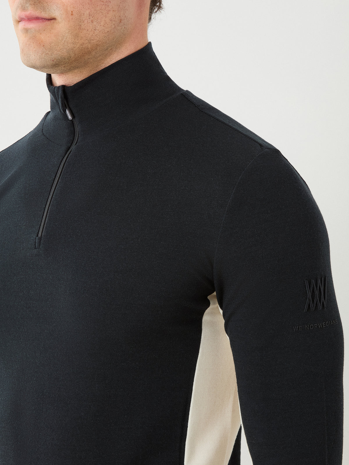 A man wears the We Norwegians Voss ZipUp Men, a black, long-sleeve quarter-zip shirt with a high collar. The shirt features activewear-inspired color blocking with white side panels and “XXXV We Norwegians” embroidered on the left sleeve.