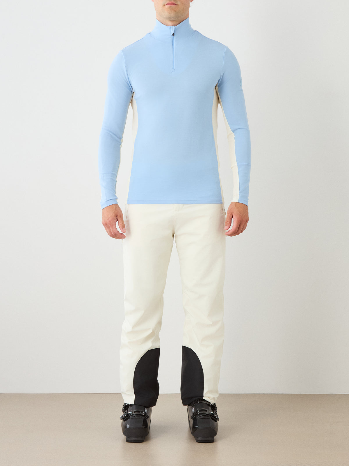 A person stands facing forward, wearing the We Norwegians Voss ZipUp Men in light blue, paired with white ski base layer pants featuring black patches on the lower legs and black ski boots, against a neutral background.