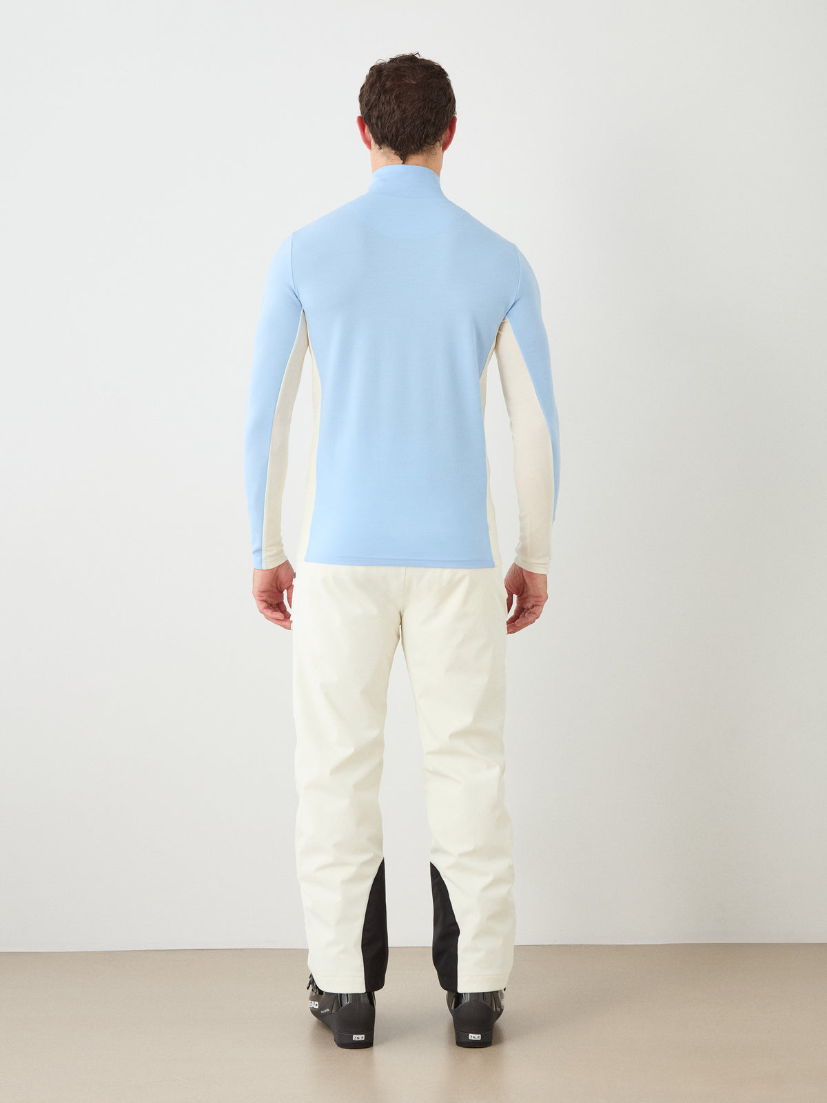 A person stands facing away against a plain background, wearing the We Norwegians Voss ZipUp Men—a light blue and cream color block jacket—layered over a cream ski base layer, paired with cream pants featuring black accents and black shoes.