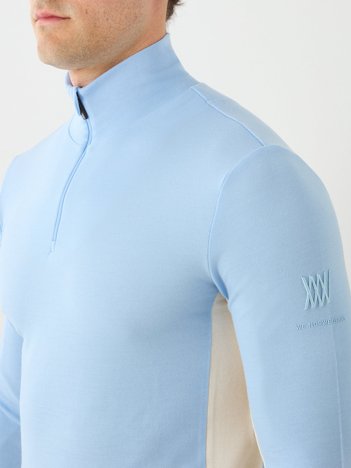 A person is pictured from the shoulders to just above the mouth, wearing the We Norwegians Voss ZipUp Men in light blue. This athletic top features white side panels and an embroidered sleeve logo—making it a perfect addition to any ski wardrobe.