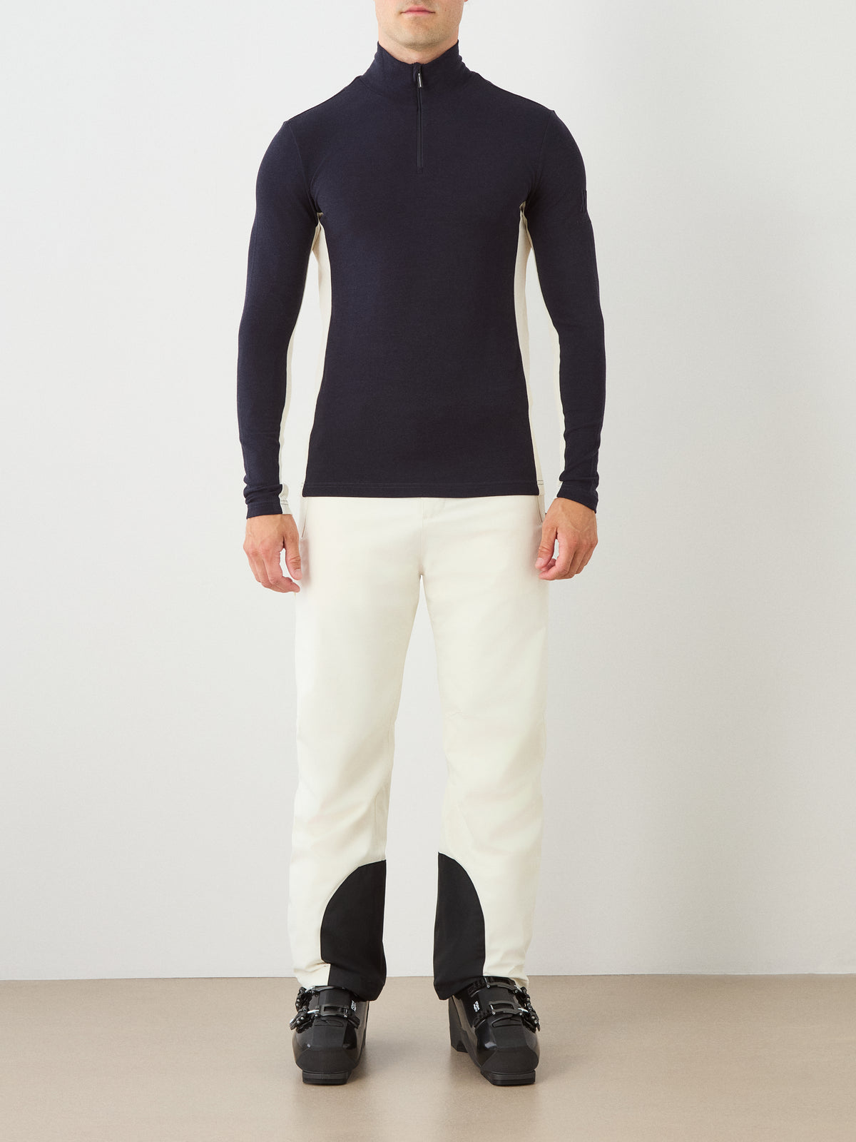 A person stands against a plain wall, wearing the We Norwegians Voss ZipUp Men in black, white pants with black patches at the ankles, and black ski boots.