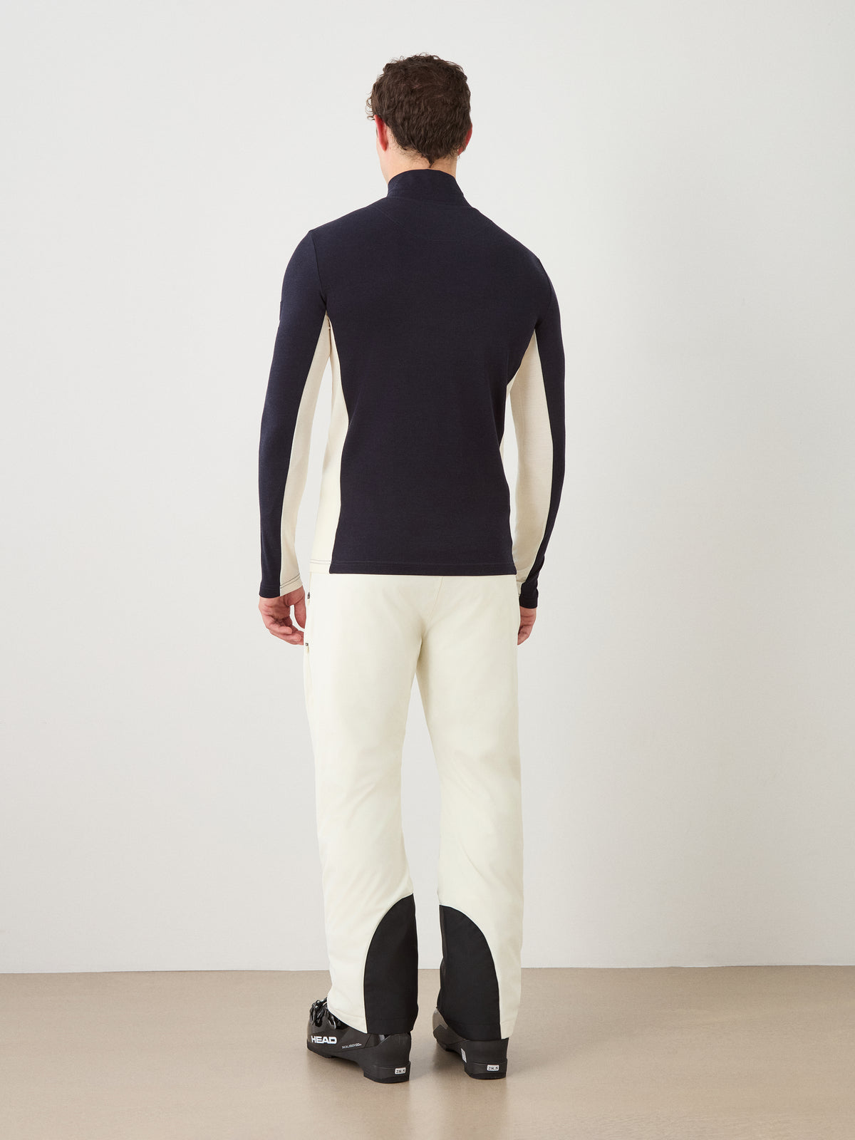 A man stands facing away against a plain light background, wearing the We Norwegians Voss ZipUp Men in black and white pants featuring black calf patches.