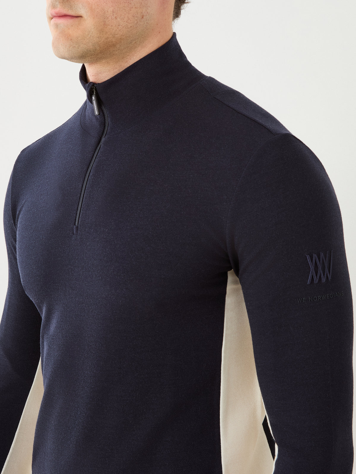 A person is shown from the shoulders to mid-torso, standing against a plain white background, wearing the We Norwegians Voss ZipUp Men—a navy blue merino wool athletic top with beige side panels.