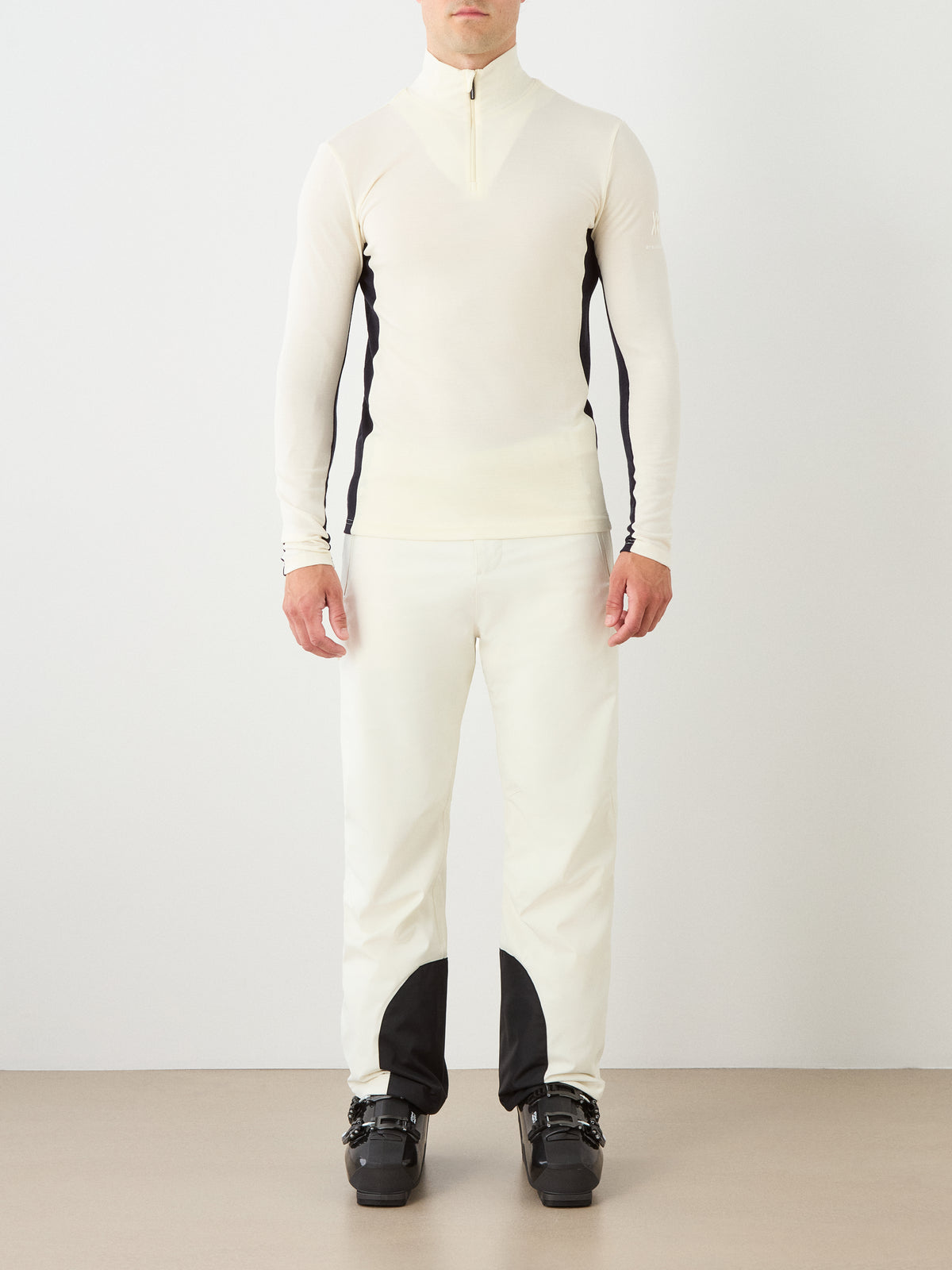 A man wearing the We Norwegians Voss ZipUp Men in cream with matching cream pants and black ski boots stands against a plain background, facing forward.