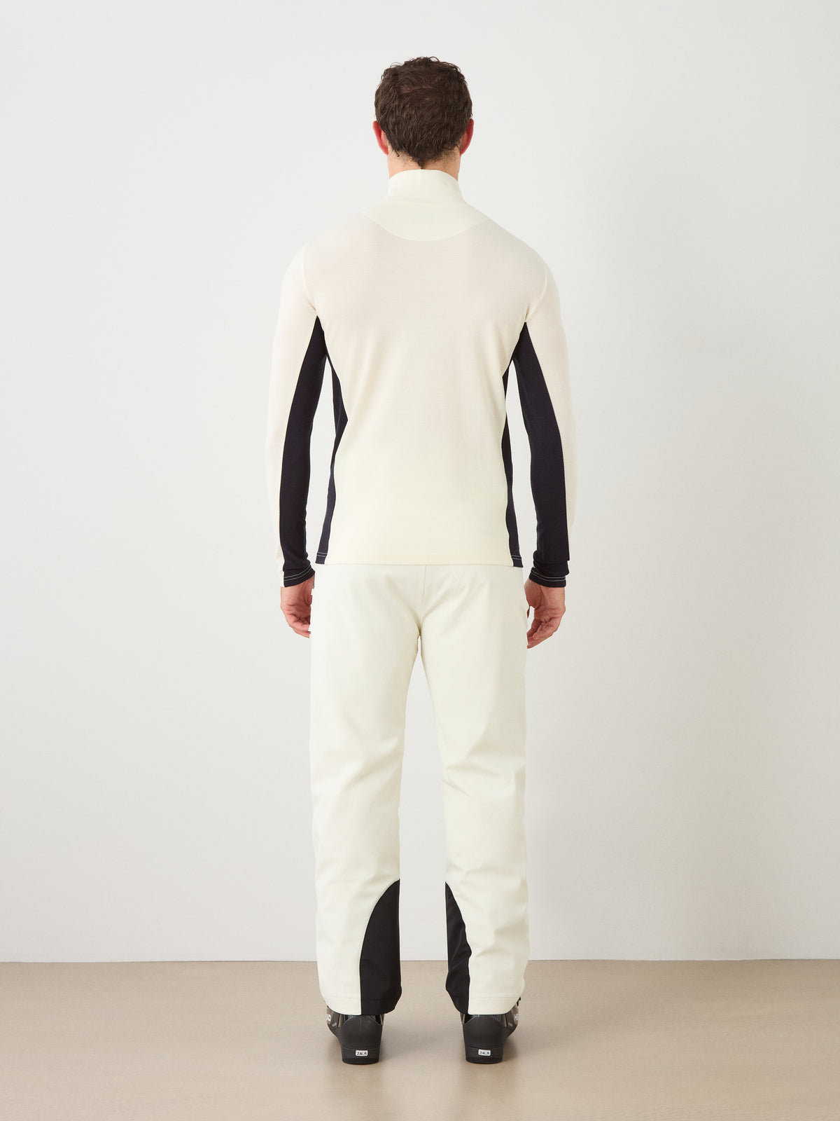 A man stands facing away, wearing the We Norwegians Voss ZipUp Men—a white long-sleeve top with black side panels inspired by classic color block jackets—paired with matching white pants featuring black accents at the ankles, set against a plain background.