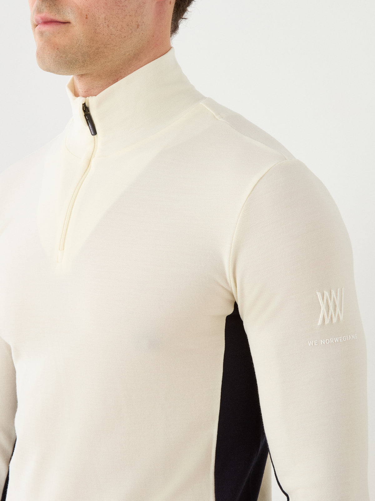 A person wearing the We Norwegians Voss ZipUp Men in cream with black side panels, styled as a sleek ski base layer. The visible sleeve displays a small white logo and “WE NORWEGIANS” text, with only the shoulders and torso shown.