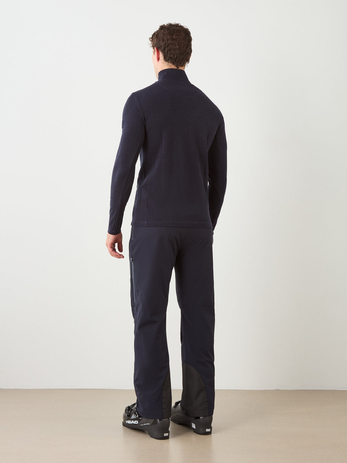 A man with short curly hair, wearing the We Norwegians Ski ZipUp Men jacket with matching pants and black ski boots, stands indoors facing away from the camera against a plain white wall.