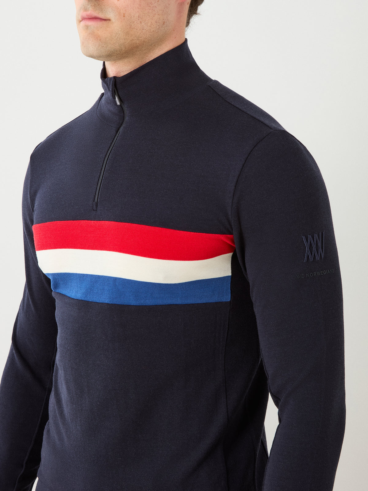 A man wears the We Norwegians Ski ZipUp Men sweater in navy, featuring red, white, and blue horizontal stripes across the chest and an embroidered logo on the left sleeve, blending retro-inspired ski wear style with modern comfort.