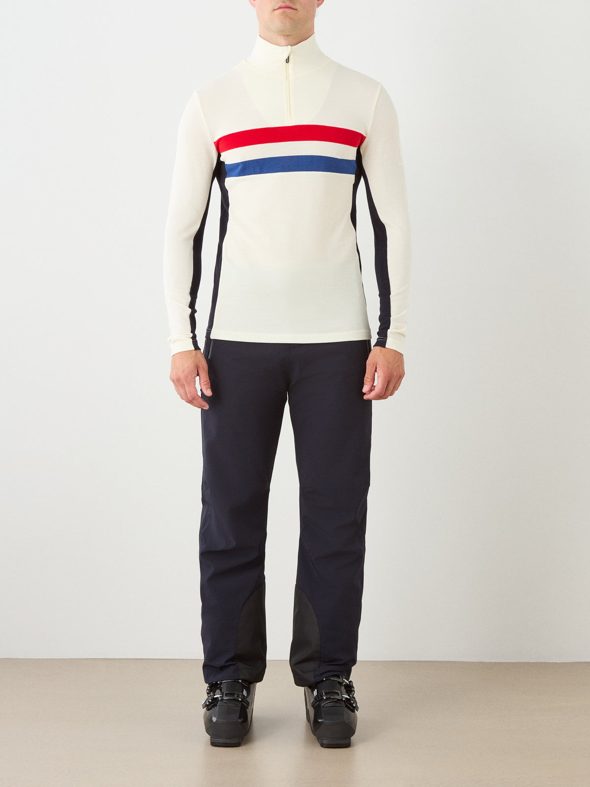 A person stands facing forward, wearing a retro-inspired ski ensemble featuring the We Norwegians Ski ZipUp Men long-sleeve top in white with red and blue stripes, paired with dark pants and black ski boots against a plain background.