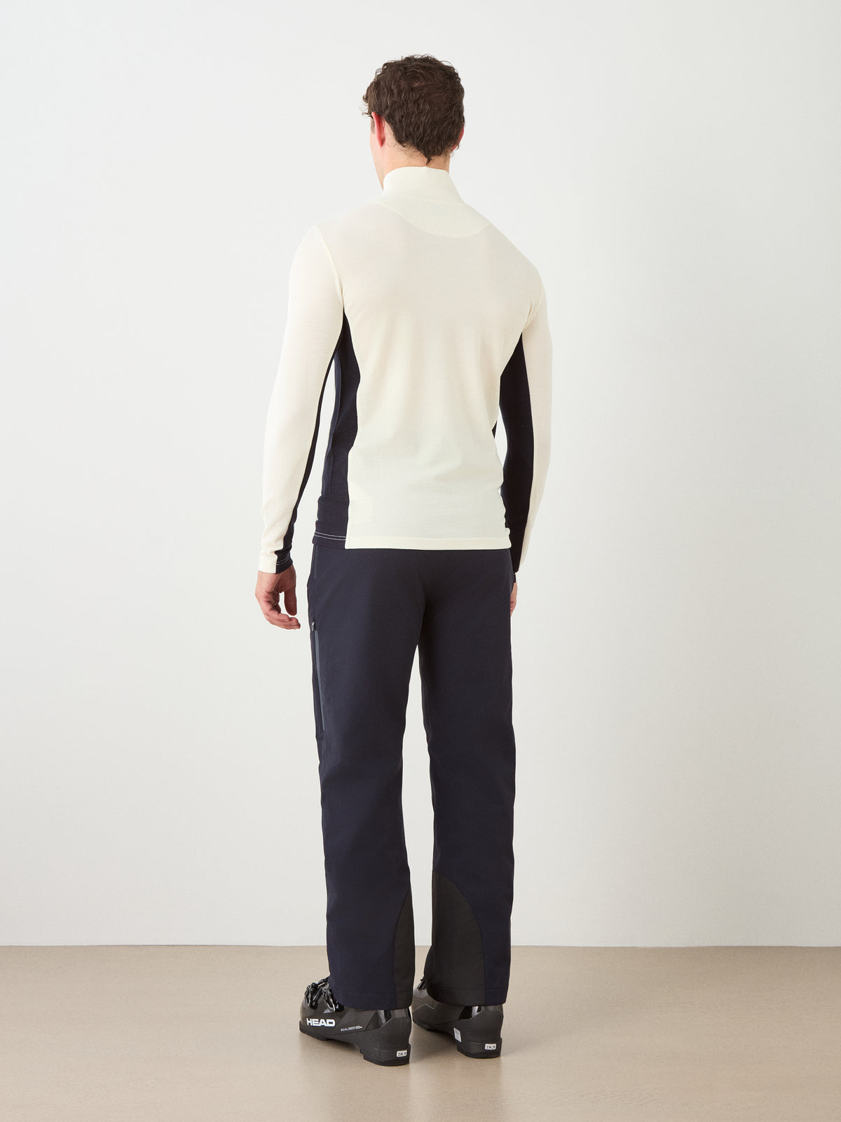 A person stands facing away, wearing the We Norwegians Ski ZipUp Men cream merino wool long-sleeve base layer top and dark athletic pants with black ski boots, against a plain white wall and beige floor.