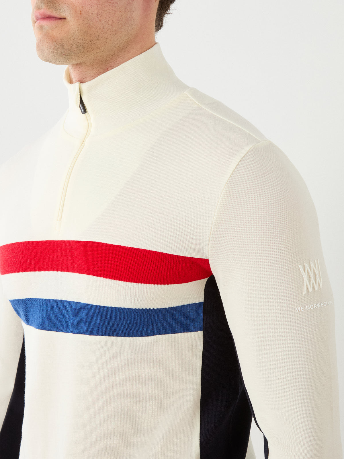 A man wearing the We Norwegians Ski ZipUp Men sweater in cream, featuring red and blue horizontal stripes across the chest and a logo on the sleeve, styled in retro-inspired ski wear, stands against a plain white background.