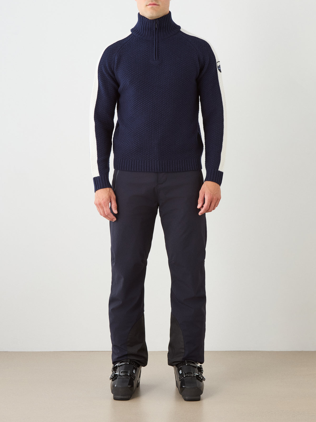 A person standing indoors against a plain light background is wearing the We Norwegians Kikut ZipUp Sweater Men in navy blue, featuring white sleeve stripes, paired with dark ski pants and black ski boots.