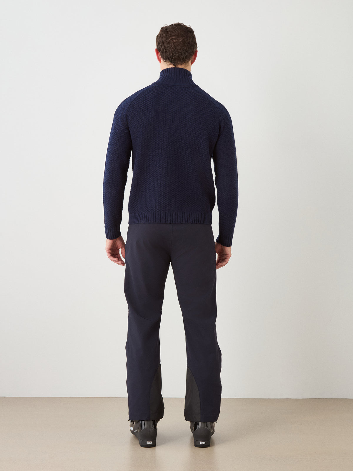 A man stands with his back to the camera, wearing the We Norwegians Kikut ZipUp Sweater Men—a classic merino wool sweater in dark blue—paired with black pants and black boots against a plain white wall and beige floor.