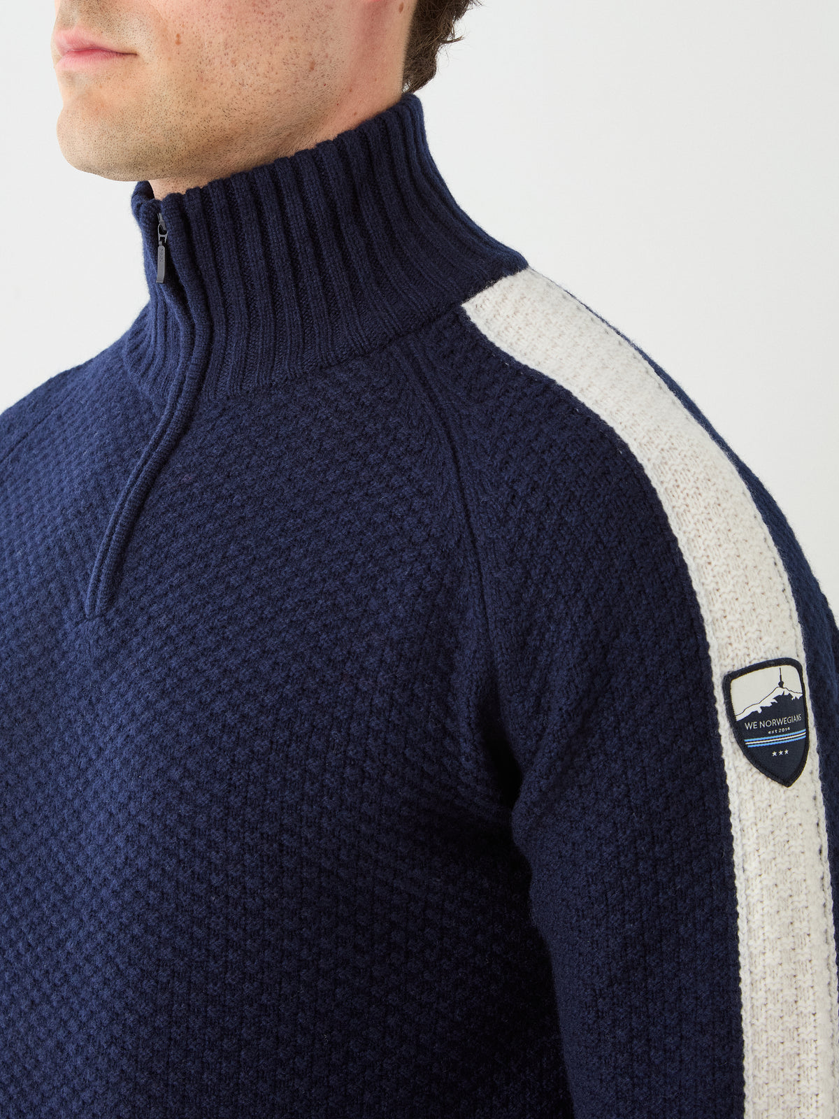 Man wearing the Kikut ZipUp Sweater Men by We Norwegians—a navy blue textured merino wool sweater with a zip-up collar, white shoulder stripe, and signature badge on the upper arm—ideal for classic après-ski style.