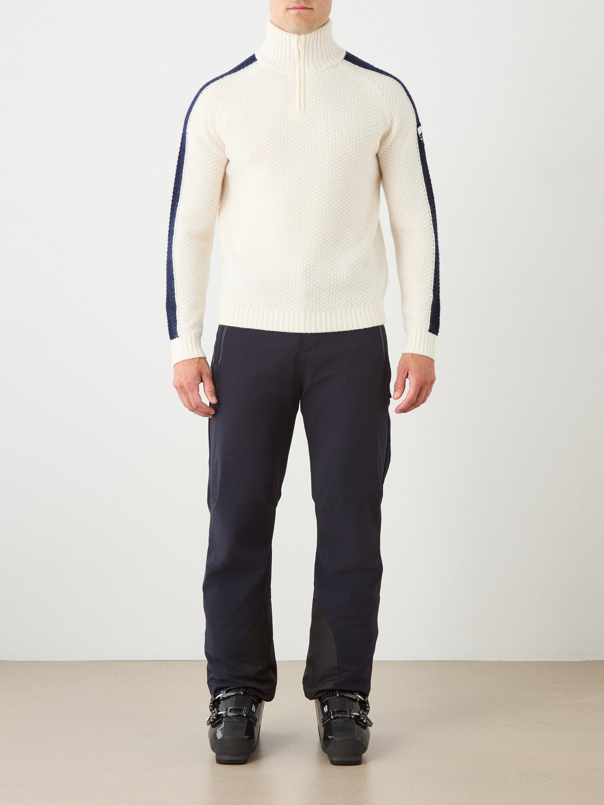 A person stands indoors wearing the We Norwegians Kikut ZipUp Sweater Men in white with navy stripes on the sleeves, paired with dark pants and black ski boots; their hands are relaxed by their sides, and their face is not visible.