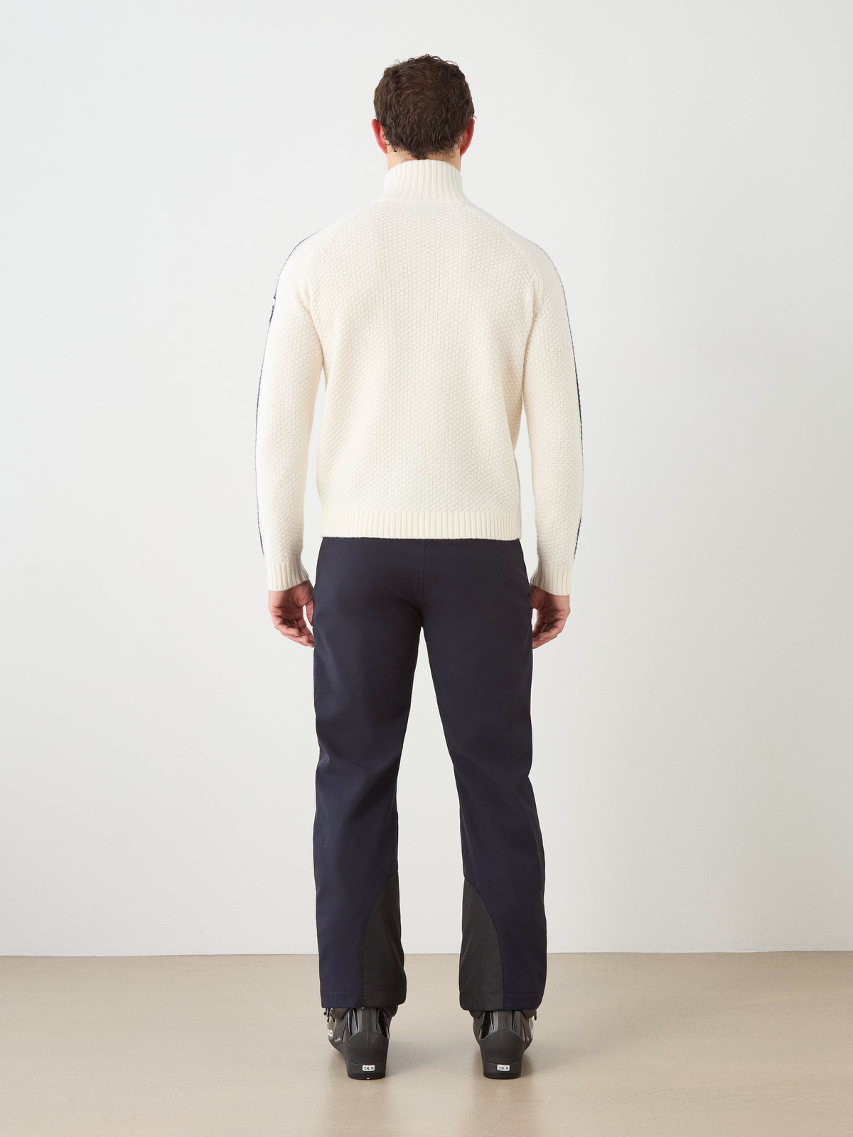 A person wearing the We Norwegians Kikut ZipUp Sweater for men in white, paired with dark pants and black boots, stands facing a white wall with their back to the camera.