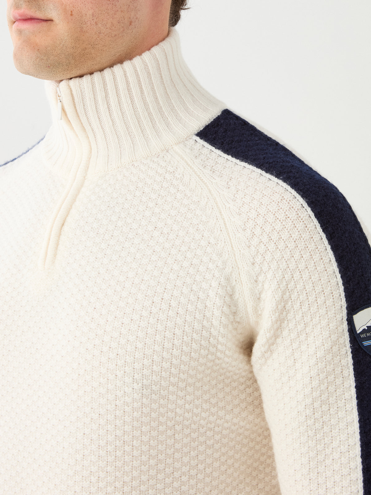 A person is seen wearing the We Norwegians Kikut ZipUp Sweater Men, a cream-colored merino wool sweater featuring a high collar, navy shoulder stripes, and a small logo patch on the upper arm. Only the lower half of their face and torso are visible.