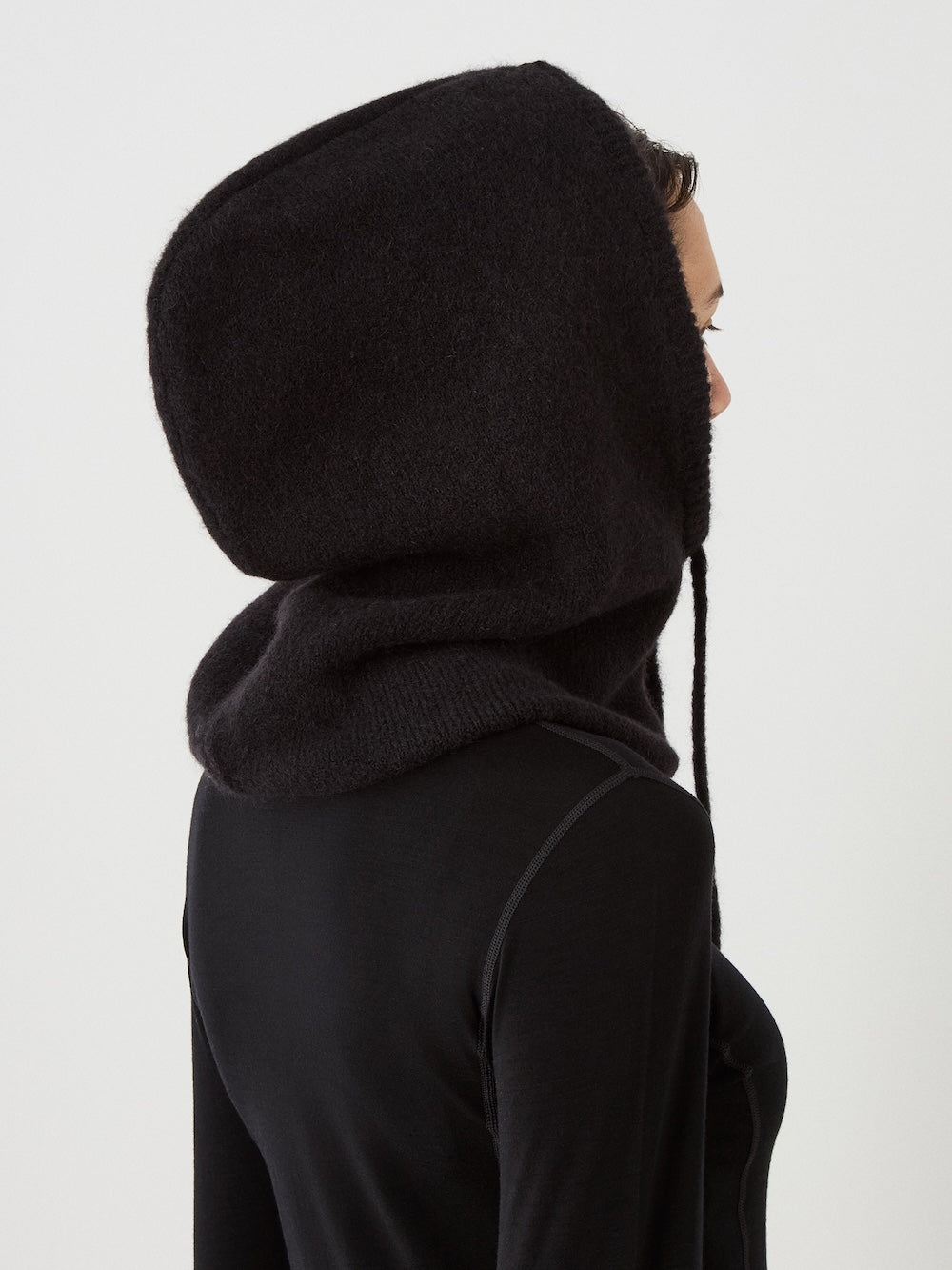 A person is wearing the We Norwegians Norefjell Hood in black, a large, cosy winter hood that covers the head and neck. The side-view image features a plain white background.