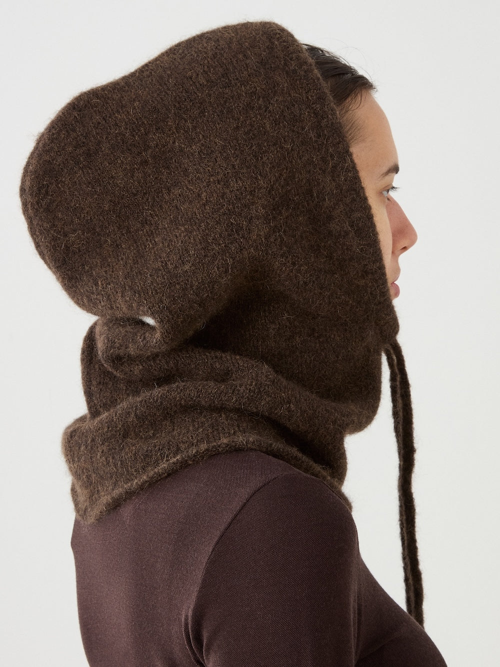 A person in profile wears the Norefjell Hood by We Norwegians—a large, textured alpaca wool piece made of thick, fuzzy material—styled as both a hood and scarf over a brown top against a plain light background, creating an effortlessly chic winter accessory.