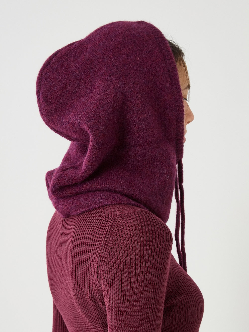 Wearing the We Norwegians Norefjell Hood, a person in a maroon ribbed sweater and matching cosy balaclava faces sideways against a plain, light background.