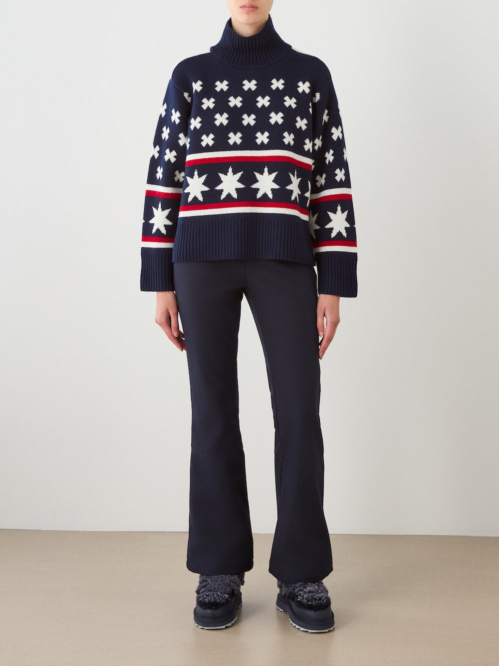 A person wearing the We Norwegians EMU x WN Sweater Women—a navy sweater featuring white star and cross patterns—paired with navy flared pants and dark boots, standing against a plain background.