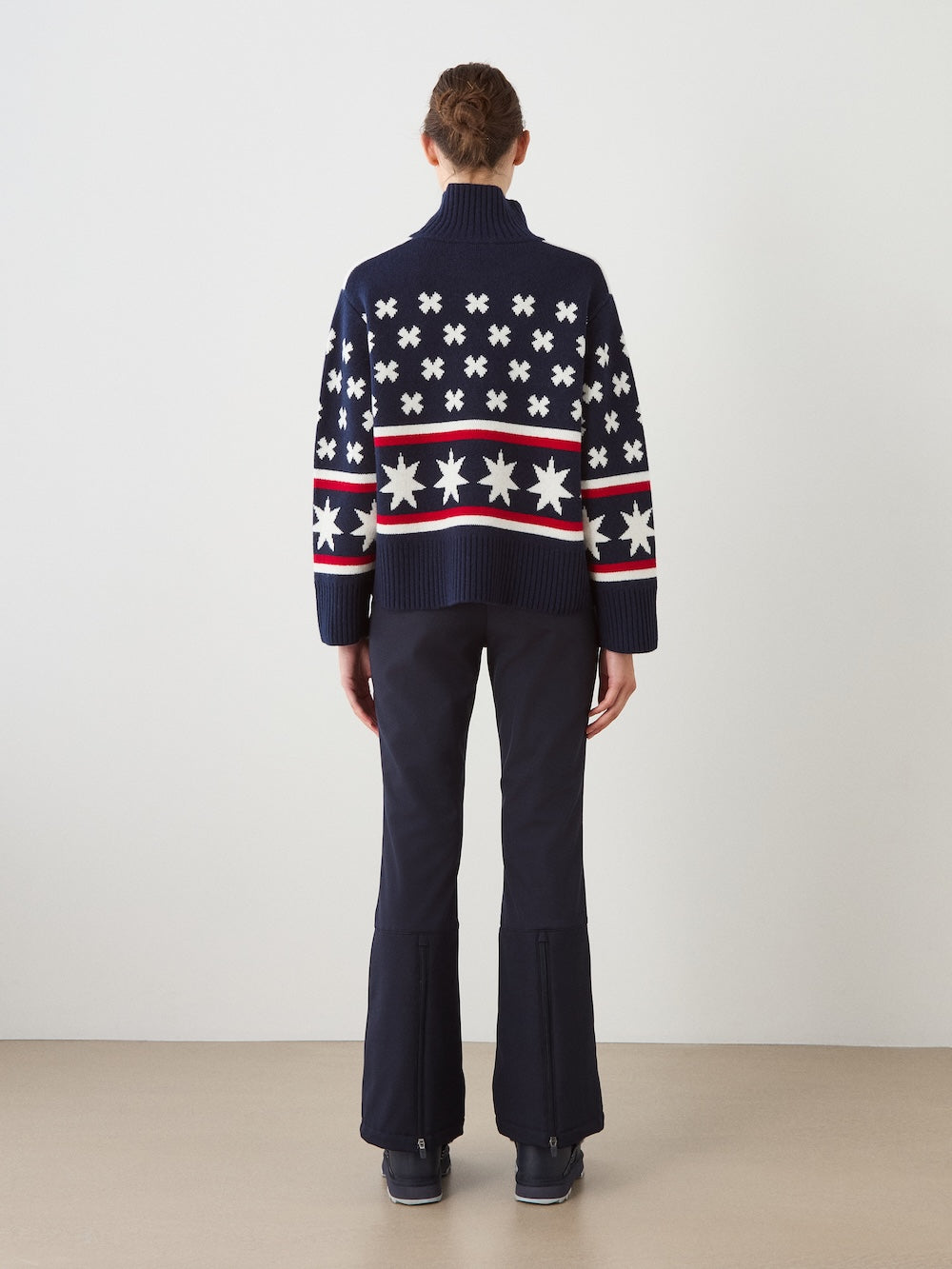 A woman stands with her back to the camera, wearing the We Norwegians EMU x WN Sweater—an oversized navy ski sweater featuring white star and cross patterns—paired with dark flared pants and black shoes, set against a plain light background.