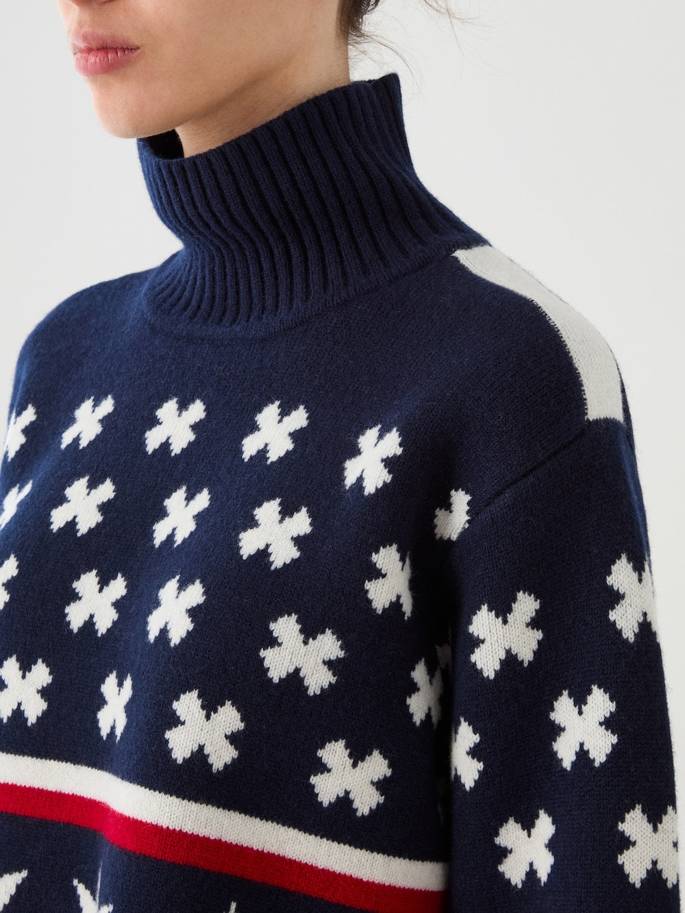A person is shown wearing the We Norwegians EMU x WN Sweater Women, a navy blue turtleneck featuring white clover-like patterns, a distinctive white shoulder detail, and a bold red and white horizontal stripe. Only the lower half of their face is visible.