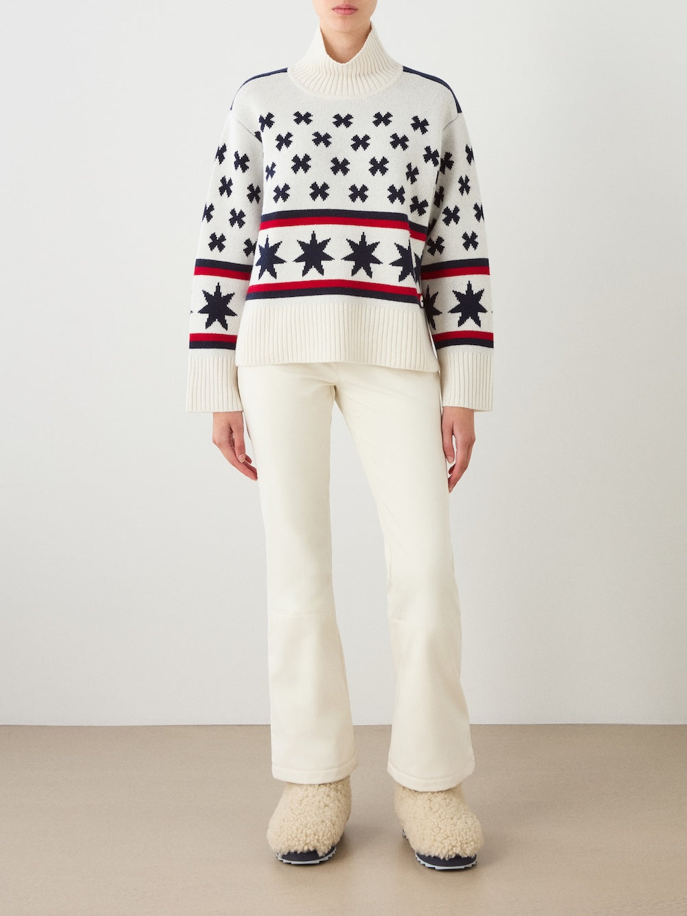 A person wearing the We Norwegians EMU x WN Sweater Women—a white merino wool ski sweater with black and red star patterns—paired with white pants and fuzzy cream slippers, standing against a plain background.
